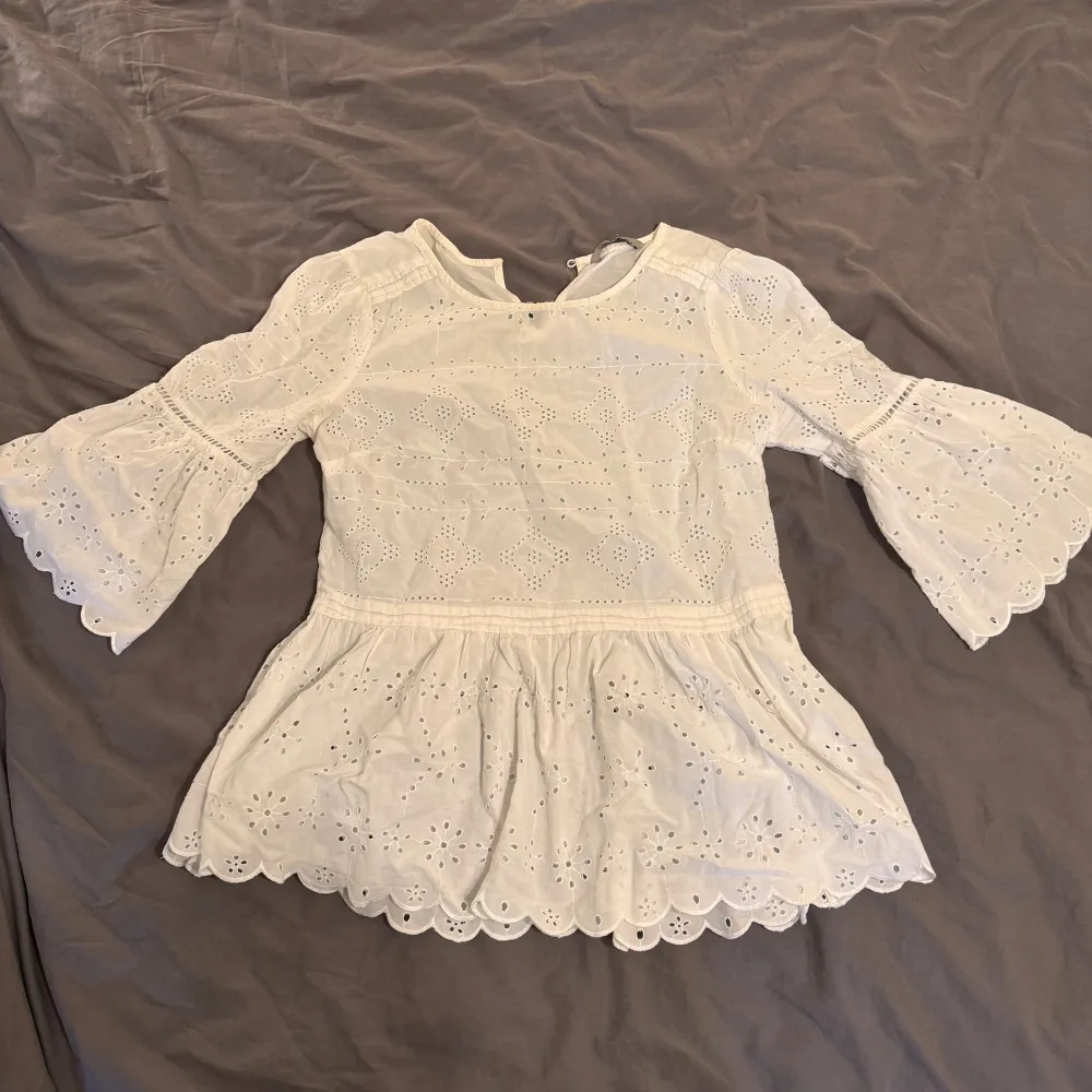 En vit blus med spetsdetaljer och broderade mönster. Blusen har trekvartsärmar och en volangkant vid ärmslut och nederkant. Den är tillverkad i ett lätt och luftigt material, perfekt för varmare väder.. Puserot.