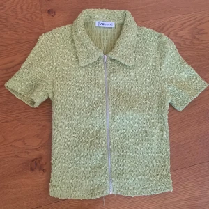 Grön kortärmad topp med dragkedja - En grön kortärmad topp från new yorker med en texturerad yta och dragkedja framtill. Toppen har en klassisk krage och är tillverkad i ett bekvämt material.