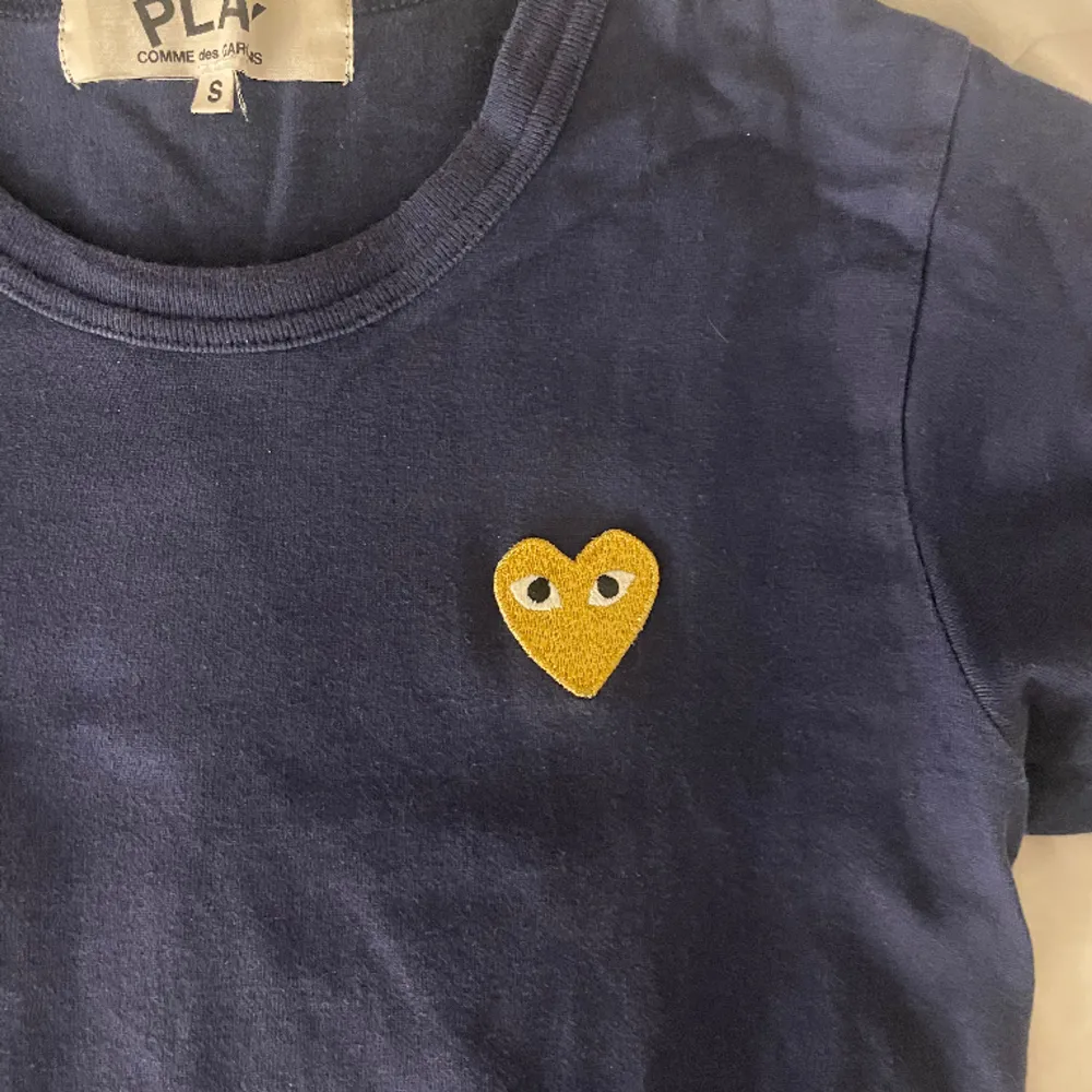 Jättefin T-Shirt från Comme des Garçons. T-shirten är marinblå med ett guldigt hjärta. Tröjan är i bra skick o använd få gånger. Skriv för fler bilder!. T-shirts.