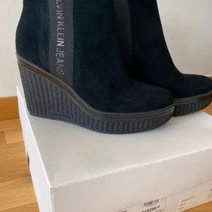 Svarta kilklackade boots från Calvin Klein Jeans - Ett par svarta boots från Calvin Klein Jeans med kilklack. Skorna är tillverkade i mocka och har en robust sula med mönster för bättre grepp. De har en elegant design med Calvin Klein Jeans-logotypen på sidan.