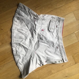 Vit kjol från Desigual - En beige kjol med en ficka bak och dekorativa sömmar. Kjolen har en knappdetalj på fickan och en broderad logotyp på baksidan.