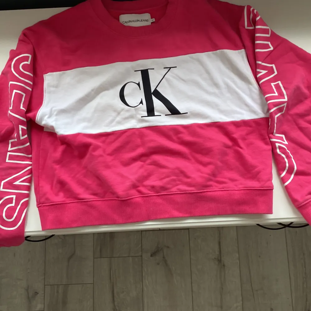 En stilren och bekväm rosa tröja från Calvin Klein Jeans med en vit panel över bröstet och stora bokstäver 'CK' tryckta i svart. Tröjan har långa ärmar med texten 'CALVIN' på ena ärmen och 'JEANS' på den andra. Ribbstickade muddar vid ärmslut och nederkant.. Tröjor & Koftor.