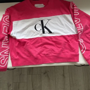 Rosa och vit tröja från Calvin Klein Jeans - En stilren och bekväm rosa tröja från Calvin Klein Jeans med en vit panel över bröstet och stora bokstäver 'CK' tryckta i svart. Tröjan har långa ärmar med texten 'CALVIN' på ena ärmen och 'JEANS' på den andra. Ribbstickade muddar vid ärmslut och nederkant.