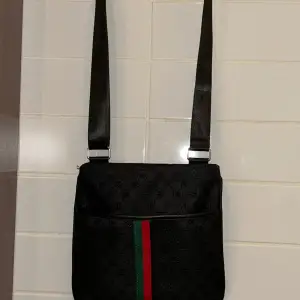 Väskan är en snygg Gucci väska.   Tyget på både fram och baksidan har loggan i svart. Framsidan har också det  klassiska grönröda bandet.  Axelremmen är justerbar, så väskan passar alla.   På framsidan finns en stor ficka med magnetknapp.  På baksida