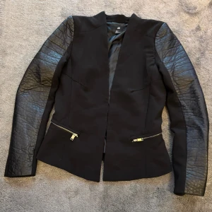 Svart jacka med läderärmar från H&M - En elegant svart kavaj/jacka från H&M med quiltade läderärmar. Jackan har en figursydd passform och två dragkedjefickor framtill. Den är perfekt för höst- och vårsäsongen.