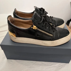Guiseppe Zanotti -  Snygga och exklusiva sneakers från Giuseppe Zanotti i svart läder med gulddetaljer. Skorna har en stilren design med guldfärgade blixtlås på sidorna och en guldig metalldetalj på hälen, vilket ger dem en lyxig touch.Perfekta för dig som vill ha en stilren och lyxig sneaker som passar både till vardags och fest. Hör av dig vid intresse!