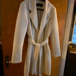 Vårkappa från Vero Moda  Strl L  - Tunn cremevit Vårkappa med knytskärp från Vero Moda. Figursydd. Material 100% polyester.  Har endast använt den ett fåtal gånger, då jag inte trivdes i den och nu är den för liten. 