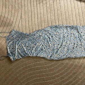 Blommmig klänning - Blå klänning med blommigt mönster. Helt i nyskick. Klänningen går att göra kortare eller längre beroende på vad man föredrar med hjälp av banden på sidorna. 