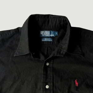 Ralph Lauren - Linneskjorta - Storlek S - VARAN KAN KÖPAS DIREKT PÅ VÅR HEMSIDA, ettmarke.se MÄRKE: Ralph Lauren. FÄRG: Svart med röd logga. MATERIAL: 55% linne, 45% bomull. STORLEK: S. AXELBREDD (SÖM TILL SÖM RAKT ÖVER): 51cm. TOTALLÄNGD (FRÅN KRAGEN): 68 cm. BREDD (PLATT MÅTT): 59 cm. 