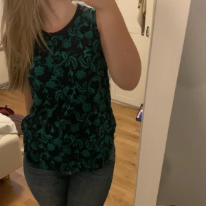 Grönt blommigt linne - Ett superfint lite längre somrigt linne. Mörk blå med grönt blommigt mönster. En super söt rosett vid ena axeln. Säljer för att den tyvärr inte kommer till användning. 