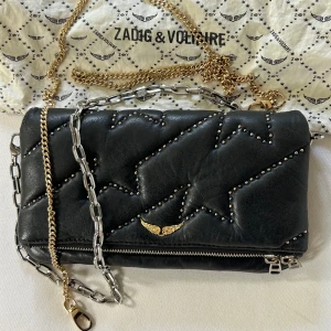 Zadig & Voltaire väska  - En ikonisk väska med dragkedja från zadig & Voltaire. Den är tillverkad i 100% Koskinn och har ”nitar” på sig.  26 cm bred och 16 cm hög.  Den har två axelband i olika längd och färg (går att ta av).  Väskan är väl använd men väldigt försiktigt.  