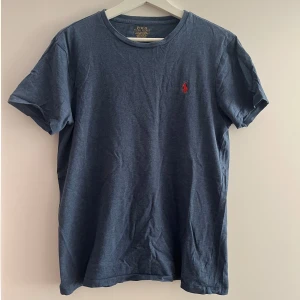 Blå t-shirt från Ralph Lauren - En blå t-shirt från Ralph Lauren med korta ärmar och en liten röd broderad logotyp på bröstet. T-shirten har en rund halsringning och är tillverkad i ett mjukt material.