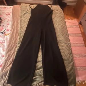 Svart långklänning med smala axelband - En elegant svart byxdress med smala axelband. byxdressen har en åtsittande överdel och vida byxben, vilket ger en sofistikerad och stilren look. 