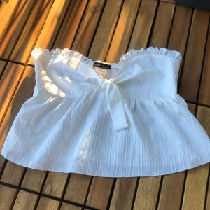 Vit tubtopp från Shein - En vit tubtopp från Shein med en söt rosett framtill. Toppen har en elastisk överdel och en volangdetalj vid fållen. Materialet är lätt och luftigt, perfekt för varma sommardagar.