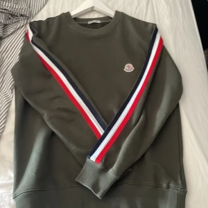Moncler tröja - En grön moncler tröja med röd,vit,blått mönster längs ärmarna. Har fått den av en kompis och vet därför inte om den är äkta. Lapparna är bortklippta.  Hör av er om fler bilder eller frågor. Pris-1100