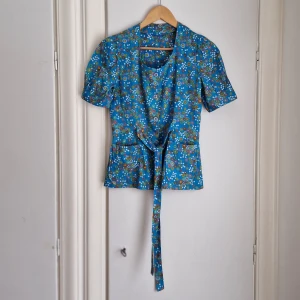 Blommigt Mönstrad retroblus  - Rolig blus med u-ringning och knytband i midjan.  Klarblå grund, med flerfärgat blomster. En riktig karamell!  Uppskattas till stl XS/S/36. Bystmått: 40 cm x 2.  Syntetmaterial, möjligtvis bomullsblandning. 