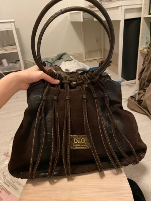 Dolce Gabbana väska  - dolce Gabbana väska, äkta mocka men osäker om äkta märke. I väl använt skick men fortfarande väldigt fin. Väskan sjunker ihop lite när man bär den så bilden hänger ”löst” ovanför väskkanten, kan ändras hos skomakare