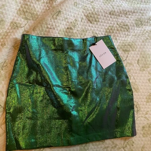 Glansig grön kjol  - Cool kjol i lite glansigt grönt material, oanvänd med prislapp kvar. Färgen är lite grönare än vad bilden visar då det var svårt att få bra ljus. Finns ingen storlek men skulle uppskatta till en xs/s
