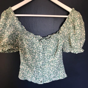 Sommartopp  - Säljer en grön topp med vita/gula blommor från Nelly i storlek 32/xs. Den har knappar fram och puffärmar. Säljer pga att den inte används💚