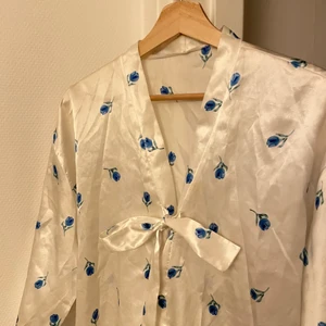 Morgonrock silke - Superskön morgonrock! Glansigt vitt material med små söta blå tulpaner(?). Går ner till knäna ungefär. Kan knytas på flera sätt så kan säker passa många men är kanske ursprungligen en storlek s/m. 