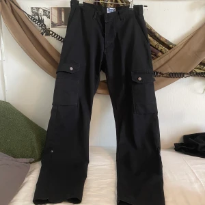 Svarta Arbets byxor - Svarta cargo byxor!! Sitter bekvämt och har många fickor, tyvär saknas en knapp (se bild)  jag är 1,60 för referens :)