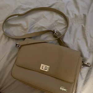 Lacoste - Säljer denna jättefina beiga Lacoste väskan! Är använd fåtal gånger, därav bra skick. Det är självklart en äkta Lacoste. Skriv om du vill ha bild på den! Nypris: 1 300kr