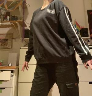 Svart sweatshirt m tryck - 65% bomull, 35% polyester. Svart sweatshirt med trycket ”BKLYN” uppe till vänster på framsidan. Två vita ränder på varje ärm. Ca 1 år gammal, hel o ren