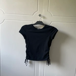 Svart t-shirt med rosetter  - En svart t-shirt med rosetter på sidorna som man knyter. Tröjan är i bra skick