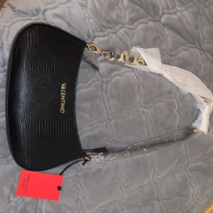 Valentino väska  - En ny valentino väska som ej har använts. Prislapp sitter kvar. Och den är inplastad. Det finns även så att man kan ha ett långt band, på ena bandet står de valentino med guldiga bokstäver. Har även kvar original kartong som väskan kom med. Köparen står för frakten 