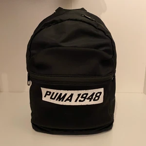 Puma 1948 ryggsäck - Svart puma ryggsäck med ett ytterfack, samt ett innerfack på insidan. I använt skick, loggan på framsidan har slitningar. Ryggsäcken är mindre än standardstorlek. H:37L:26B:13 (cm)