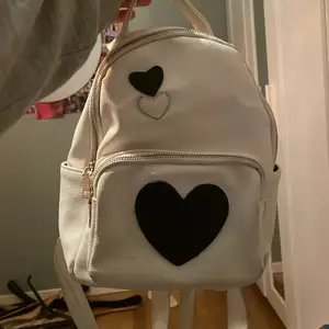 En vit ryggsäck i fake läder med svarta och vita hjärtan på köpt i Bulgarien. Två olika fack samt så är det små fack inuti väskan! Det är en liten fläck fram på väskan men inget man märker av.