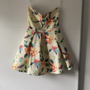 Axelbandslös klänning - Blommig klänning utan axelband. Tyllkjol under för extra volym, dragkedja på baksidan och ett stål pinne håller det lilla ”v” i ringningen snygg. Storleken är 12 vilket motsvarar 38. Kan mötas upp i Göteborg/Nyköping alternativt skicka om kund står för frakt 🌼