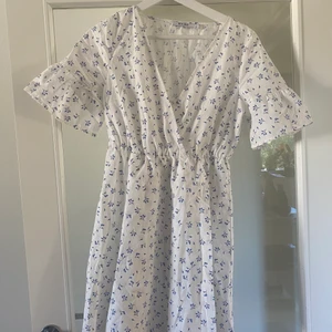 Klänning NAKD  - Supersöt kort blommig klänning från NAKD, som går i färgerna blått och vitt! Klänningen är i en omlottmodell, resor i midjan och korta volang ärmar! Använd endast vid 2 tillfällen! 🌻🌼