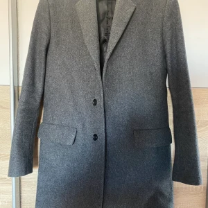 Filippa K Rhine Recycled Melton Wool Coat  - Filippa K Gray Rhine Recycled Melton Wool Coat i storlek 46.  Rock i en klassisk modell med rak passform från Filippa K. Producerad i en kraftig återvunnen ullblandning, med tunt foder, som passar utmärkt för kyliga höstdagar.  • Stängs med knappar. • Två framfickor med lock, samt en bröstficka. • Tre innerfickor. • Enkelt sprund. Ordinarie pris på careofcarl.com: 3999kr. Buren en gång för att se passform.