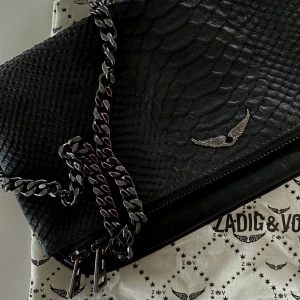 Zadig & Voltaire Rock Savage bag - En svart väska från Zadig&Voltaire i kobra liknande läder. Tillkommer två flyttbara kedjor, väskan går därför att ”carry on shoulder or across the body”. Dustbag tillkommer. Väskan har tre mindre fack och rymmer mycket. Ordinarie pris 360€. Använd men är i mycket bra skick. PRISET SOM STÅR ÄR DET SOM GÄLLER!                                  Width x Height x Depth : 27 x 13 x 2 cm.
