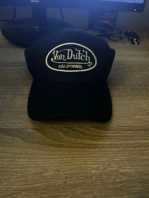Svart Von Dutch keps med mesh - Säljer en svart keps från Von Dutch med klassisk broderad logga framtill och texten 'California'. Kepsen har böjd skärm och mesh på sidorna och bak för extra ventilation. Perfekt för dig som gillar streetwear och vill ha en ikonisk accessoar. Helt ny.