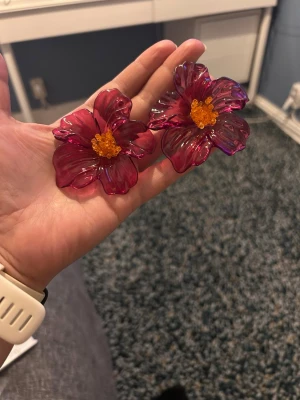 Stora blommiga örhängen i rosa plast - Säljer ett par statement örhängen formade som stora blommor i transparent rosa plast med gula detaljer i mitten. Örhängena är riktigt iögonfallande och har en cool, lekfull vibe. Perfekta för dig som vill sticka ut och addera färg till din stil.