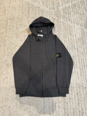 Mörkgrå hoodie från Stone Island - Snygg mörkgrå hoodie från Stone Island med dragkedja framtill och klassisk logopatch på vänster ärm. Hoodien har justerbar huva med snören, ribbade muddar och två fickor framtill. Perfekt för en avslappnad och trendig stil.