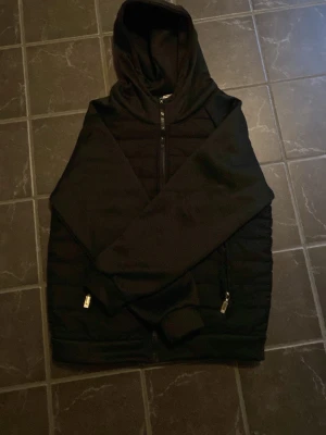 Svart cardigan med huva - Snygg svart vindjacka med dragkedja framtill och två fickor med dragkedja. Jackan har quiltade partier på kroppen och släta ärmar. Utrustad med huva för extra skydd mot vinden. Perfekt för dig som vill ha en stilren och funktionell jacka.