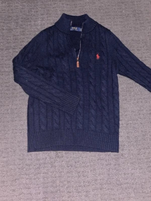 Mörkblå kabelstickad tröja Polo Ralph Lauren - Mörkblå kabelstickad tröja från Polo Ralph Lauren med halv dragkedja och röd broderad logga på bröstet. Tröjan har ribbstickade muddar och hög krage, perfekt för lager-på-lager-stil.