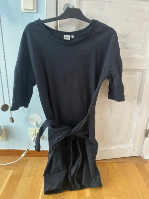 Svart klänning med knyt i midjan - En svart klänning från object i mjuk bomull med rund halsringning och knytband i midjan. Klänningen har korta ärmar och en rak, enkel siluett som ger en clean och avslappnad look. Perfekt för dig som gillar minimalistisk stil. Fläckar på den vilket syns på bilderna, osäker på om det går bort i tvätten🫶🏼