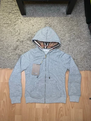 Grå zip hoodie från Burberry - Snygg grå zip hoodie från Burberry med klassiskt rutigt foder i huvan och broderad logga på bröstet. Mjuk bomullsblandning, två fickor fram och ribbade muddar. Perfekt för en chill och stilren look.