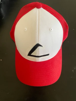 Röd och vit truckerkeps med mesh - Säljer en röd och vit truckerkeps från YUPOONG Classic med justerbar snapback. Kepsen har vit frontpanel med svart tryck och röd böjd skärm samt mesh på sidorna och bak för extra ventilation. Perfekt för dig som gillar streetwear och retrostil.