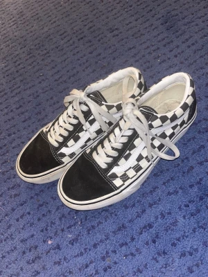 Vans Old Skool Checkerboard sneakers - Klassiska Vans Old Skool sneakers med svart och vitt checkerboard-mönster, vita snören och sula samt svart tåparti. Ikonisk sidostripes i vitt och röd Vans-logga på hälen. Ovandel i canvas och mocka för en skön och stilren look.