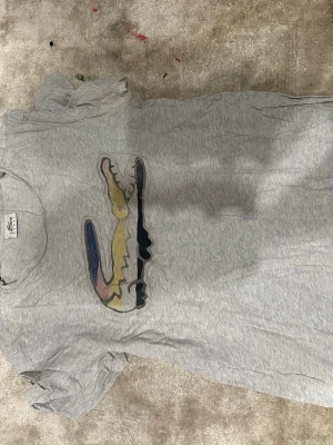 Grå Lacoste t-shirt med färgglad logga - Snygg grå t-shirt från Lacoste med den klassiska krokodilloggan i flera färger på bröstet. T-shirten har rund hals och korta ärmar, perfekt för en avslappnad stil. Tillverkad i mjuk bomull för skön komfort.