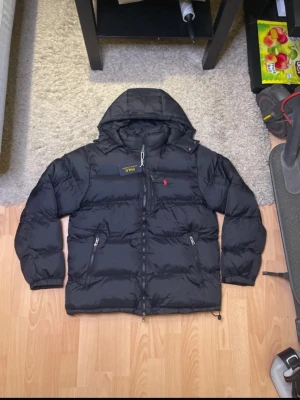 Svart dunjacka från Polo Ralph Lauren - Svart dunjacka från Polo Ralph Lauren med röd broderad logga på bröstet. Jackan har huva, två dragkedjeförsedda fickor och quiltad design. Perfekt för kalla dagar och har en klassisk pufferstil.