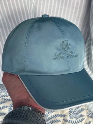 Ljusblå keps från Loro Piana - Snygg ljusblå keps från Loro Piana med broderad logga framtill. Klassisk sexpaneldesign och böjd skärm. Kepsen har en stilren look och är tillverkad i ett slätt, exklusivt material som känns lyxigt.