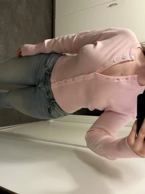 Ljusrosa ribbad kofta med knappar - Supersöt ljusrosa kofta med ribbad struktur och vita knappar hela vägen framtill. Figurnära passform och långa ärmar.