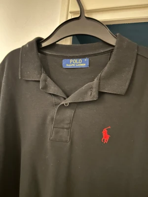 Svart pikétröja från Ralph Lauren - Svart pikétröja från Polo Ralph Lauren med klassisk krage och två knappar. Tröjan har det ikoniska röda broderade polospelar-loggan på bröstet. Tillverkad i bomull och har korta ärmar. Perfekt för en stilren och avslappnad look.