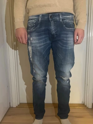 Blå slim fit jeans från Replay - Säljer ett par blå slim fit jeans med snygga slitningar och tvättade detaljer. Jeansen har fem fickor, vita kontrastsömmar och en modern passform som sitter tajt längs benen. Perfekta för en trendig och avslappnad stil. Jeans modell på jeansen är bronny. Personen på bilden är 185 cm lång 